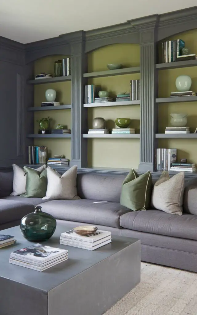 A photo of a gray and green living room with built-in bookshelves painted in alternating gray and green tones. A very cozy gray sectional sofa sits in front, styled with green and gray throw pillows. The bookshelves hold a mix of books, green ceramic pieces, and small plants. A low rectangular coffee table adds function, with a stack of magazines and a green glass vase for decoration.