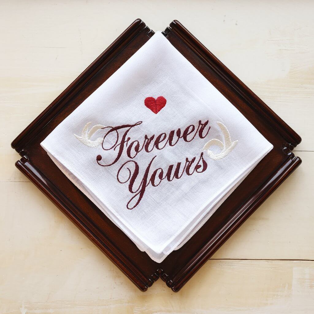 A handkerchief embroidered with a small red heart and the phrase "Forever Yours" in elegant script. It is displayed on a polished white wooden surface. The handkerchief is folded into a triangle shape. The background is clean and simple, with no additional elements.