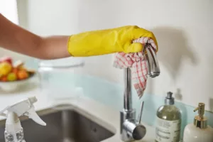 a hand wearing yellow cleaning gloves, cleaning a faucet with a soft cloth