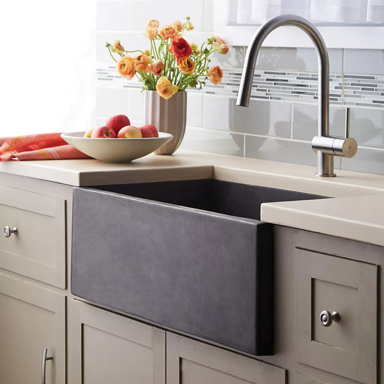 A modern kitchen featuring a stone sink integrated into a light-colored countertop, with a bowl of fresh fruits and a vase of orange flowers nearby, under the natural light shining through the window.
