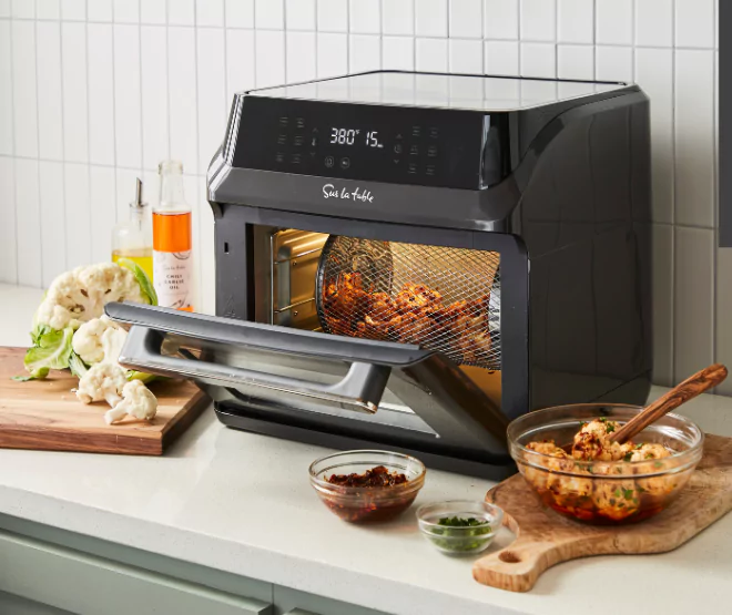 A **Sur La Table Air Fryer** with its door open, revealing food inside. Various ingredients and condiments are arranged around it, indicating the preparation of a meal. The air fryer is black, and digital controls are visible on the top, displaying the temperature and time. A bottle of oil or some liquid ingredient is to the left of the air fryer, and small bowls containing chopped vegetables and spices are in front of it. A wooden spoon rests in one of the bowls filled with a mixed ingredient, possibly a salad or marinated food item. Leafy greens and other unidentifiable ingredients are scattered around the countertop.