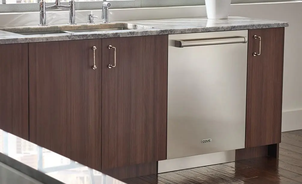 This is an image of a Cove DW2450 dishwasher. The dishwasher is integrated seamlessly into a kitchen cabinet with a dark wooden finish and a granite countertop, providing a sleek and clean look to the kitchen space. The dishwasher is built-in, blending seamlessly with the dark wooden cabinets on its sides. Above the dishwasher is a light-colored granite countertop with an undermount sink and two faucets. The cabinet doors have silver handles that match the stainless steel front of the dishwasher. The brand “COVE” is visible on the lower right corner of the dishwasher door. Natural light illuminates the scene, highlighting the textures and finishes of both wood and stone materials.