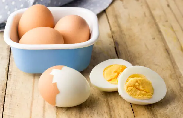 The image is a blue bowl containing four whole brown eggs, a peeled hard-boiled egg with a crack, and another hard-boiled egg cut in half revealing the cooked yolk, all placed on a wooden surface. The eggs are well-cooked and appear to be fresh. The wooden surface appears rustic and worn, and the lighting is natural and soft, illuminating the eggs and emphasizing their textures.
