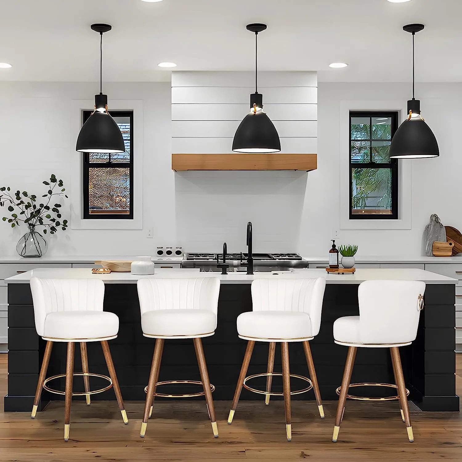 “A modern kitchen interior featuring a set of four SeekFancy 27” Swivel Bar Stools with white seats and wooden legs, positioned at a black and white kitchen island. Above the island are three black pendant lights, illuminating the space."