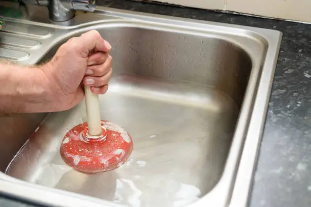 The image shows a person using a red plunger to unclog a stainless steel kitchen sink. The person’s hand is visible, gripping the handle of the plunger firmly. The sink appears to be empty and clean, with no visible debris or water. The countertop surrounding the sink is made of a dark, speckled material, possibly granite or a similar stone. In the background, there is part of another sink and what appears to be bottles of cleaning products or similar items.
