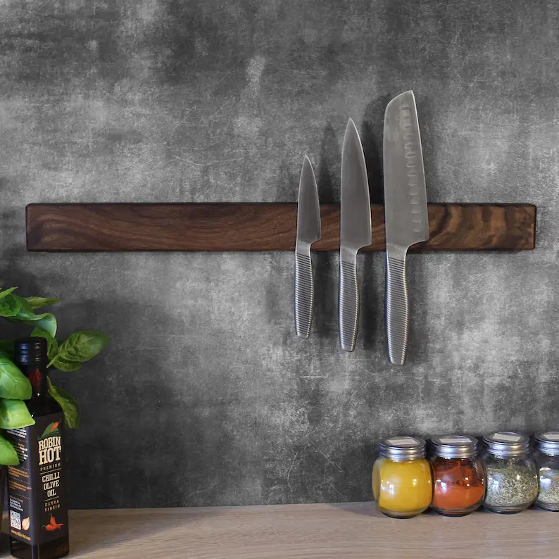 A magnetic strip with three knives attached, mounted on a dark wooden surface. Spice jars and a bottle of hot sauce are visible on the counter below.