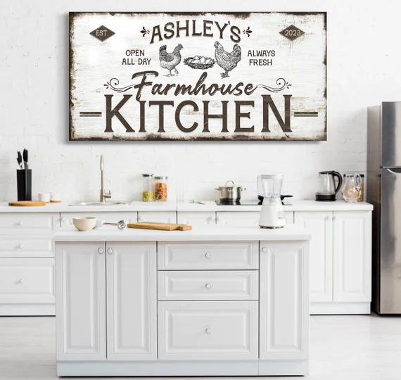The image shows a modern, well-lit kitchen with white walls and countertops. A large decorative sign with a rustic design that reads “ASHLEY’S Farmhouse KITCHEN” is hung on the wall above the countertop. The countertop is white, clean, and contains various items including a cutting board, knife block, containers, and modern appliances like a coffee maker. The cabinets below the countertop are also white with silver handles; they appear to be well-maintained and clean. To the right of the image is part of a stainless steel refrigerator. The sign has text and illustrations that read “ASHLEY’S Farmhouse KITCHEN EST 2022 OPEN ALL DAY ALWAYS FRESH”