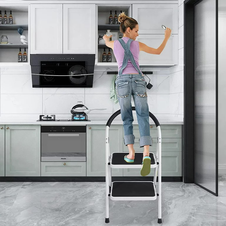 folding step stool in the kitchen with a woman stepping on it to take something in the cabinet 