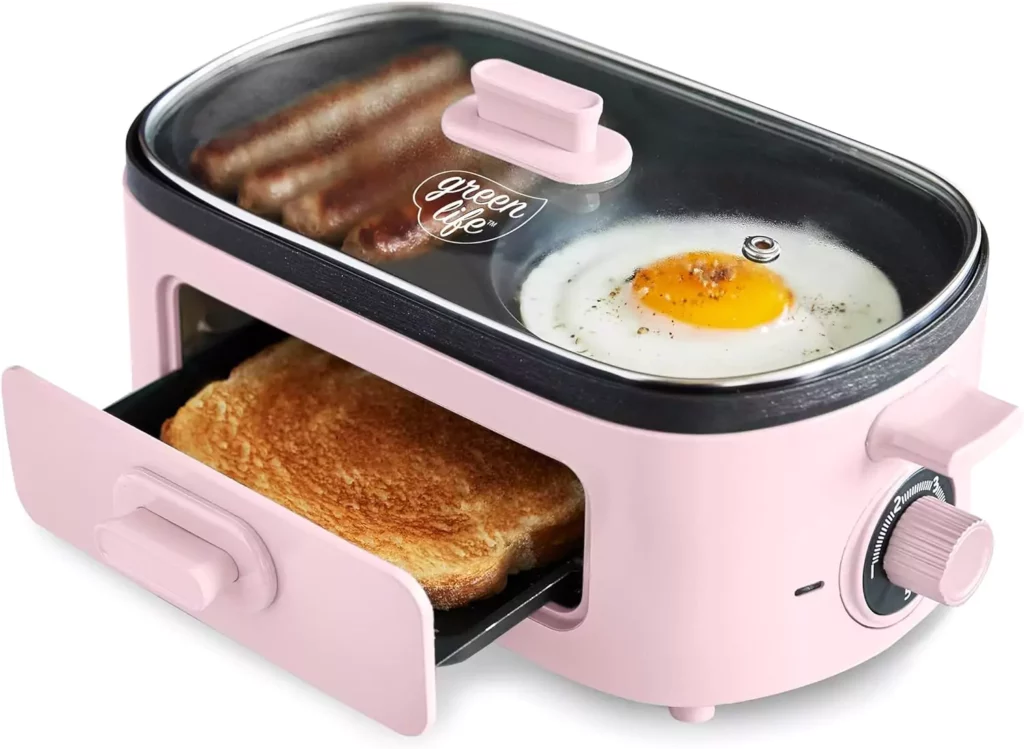 A GreenLife 3-in-1 Breakfast Maker Station in pink color, with a slice of bread being toasted in the drawer, sausages and an egg frying on the top griddle. The appliance has a round control knob on its right side for temperature adjustment. 