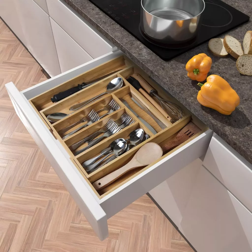 A neatly organized kitchen drawer with wooden dividers, holding a variety of utensils including spoons, forks, and knives. The drawer is part of a modern kitchen setup with a countertop where a pot and fresh bell peppers are visible.