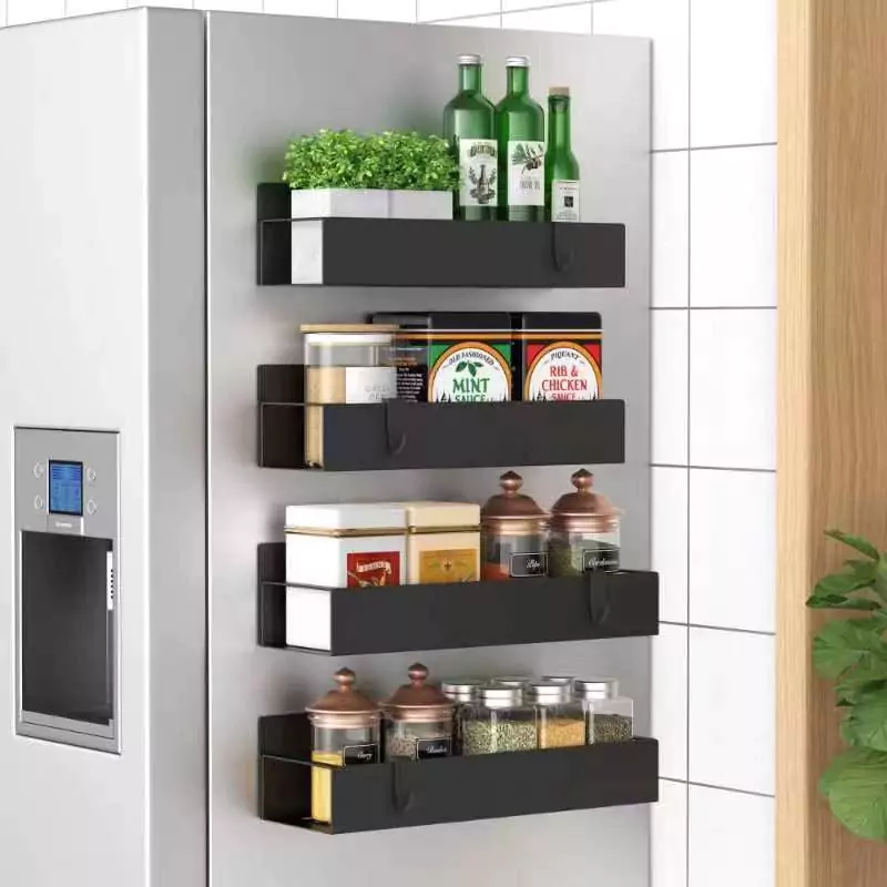 A modern kitchen setting featuring a stainless steel refrigerator with a digital screen, next to a white tiled wall and wooden panel. Three black magnetic shelves are attached to the fridge, holding an assortment of items including green plants, bottles, jars of spices, and containers.