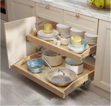 A well-organized kitchen drawer with neatly stacked plates, bowls, and pots. The upper level holds white plates, yellow bowls, and smaller white dishes, while the lower level contains silver pots with glass lids and a blue pot without a lid. The drawer is part of a light-colored kitchen cabinet with elegant handles. In the background, there are green bananas on a countertop.