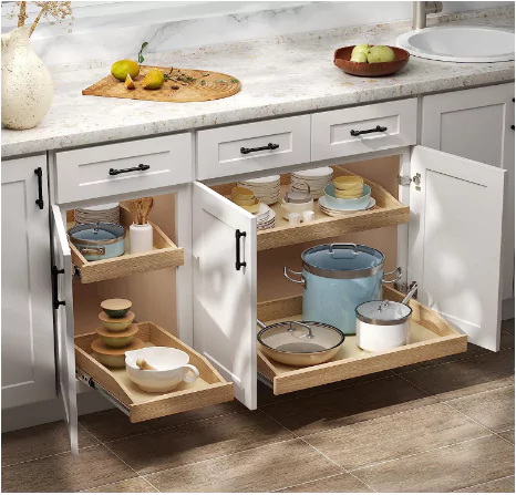 A well-organized kitchen cabinet with pull-out shelves, stocked with various dishes, pots, and utensils. The upper shelf contains plates, bowls, and cups, while the lower shelf holds a large blue pot and other cooking utensils. On the countertop above the cabinets are wooden trays with fruits like bananas and apples. The kitchen floor appears to be made of tiled or polished stone.