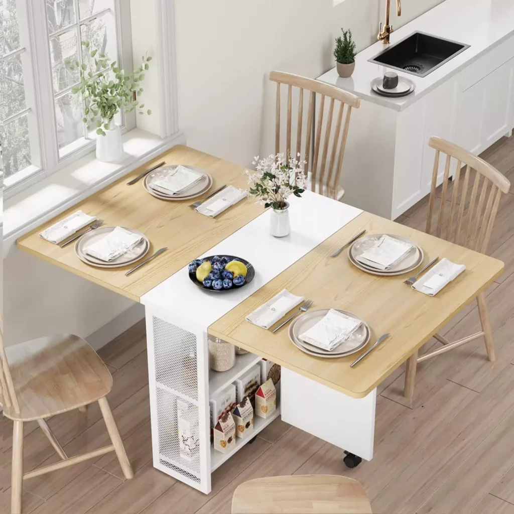 A bright and cozy dining area with a modern wooden table set for four, featuring a mix of chairs and stools. Fresh flowers and a bowl of fruit adorn the table, and the room is adjacent to a well-equipped kitchenette.
