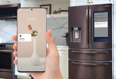 A person’s hand holding a smartphone displaying an augmented reality (AR) application that overlays a virtual vase with flowers on a real-time image of a kitchen, with a modern refrigerator in the background.