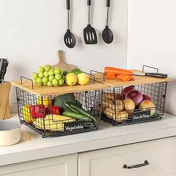 A well-organized kitchen countertop with wire mesh storage baskets containing fresh fruits and vegetables. Utensils hang on the wall in the background. The countertop appears to be made of a light-colored material, possibly quartz or marble.
