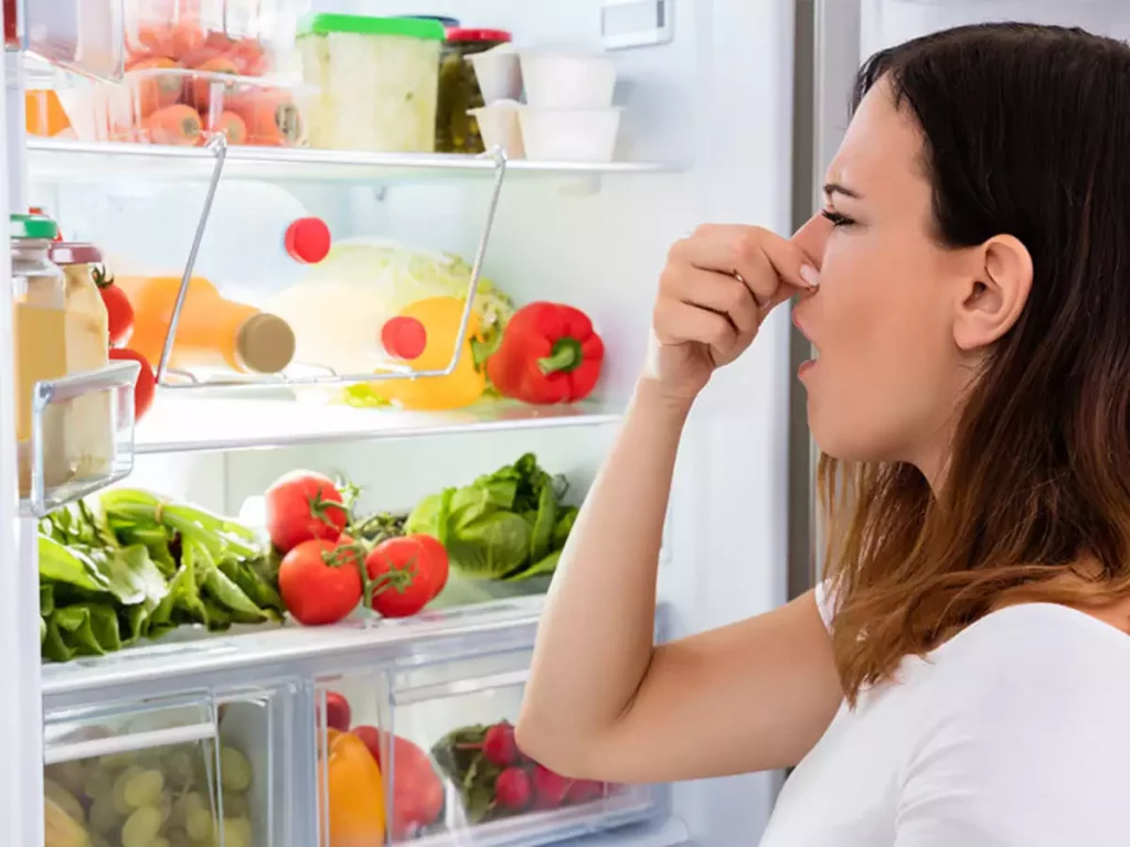 An individual with their face obscured is peering into a well-stocked refrigerator filled with various fresh produce and other food items. 