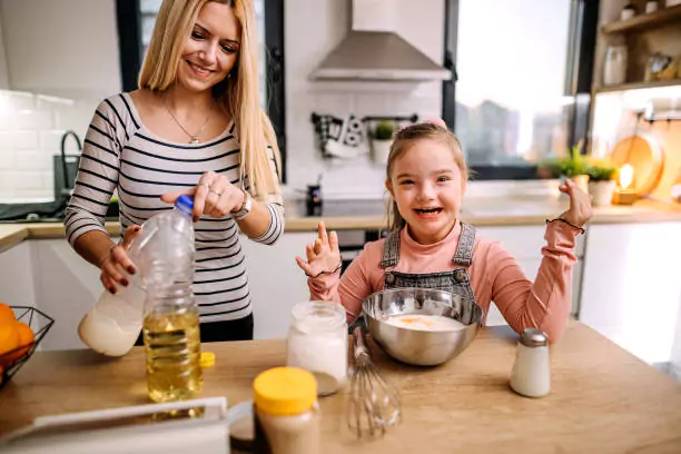 Two individuals in a modern kitchen, engaging in baking. One is pouring oil into a mixing bowl while the other excitedly raises their hands.
