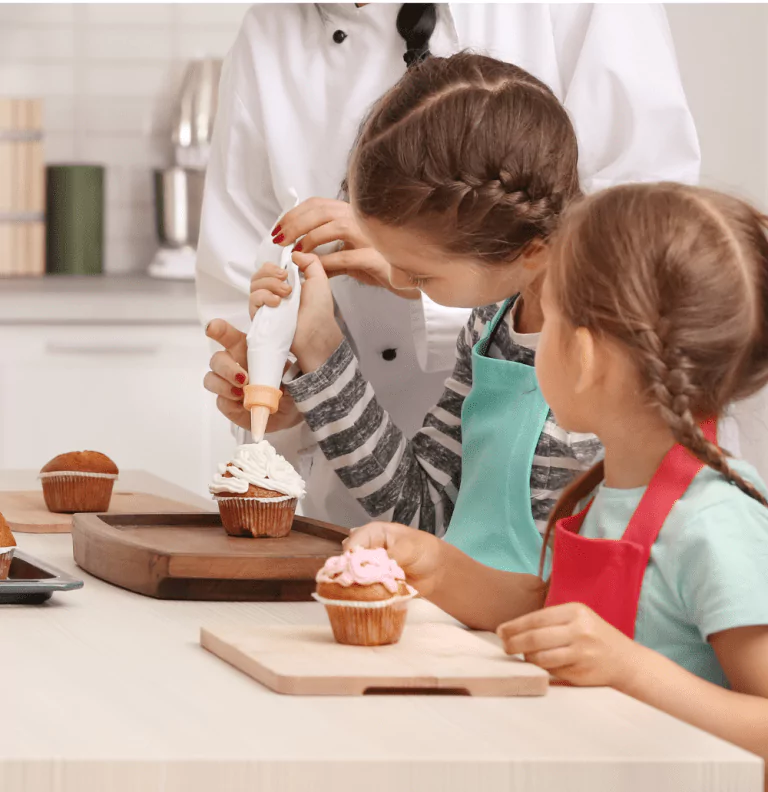 Two children decorating cupcakes with icing in a kitchen, under the supervision of an adult in chef attire.