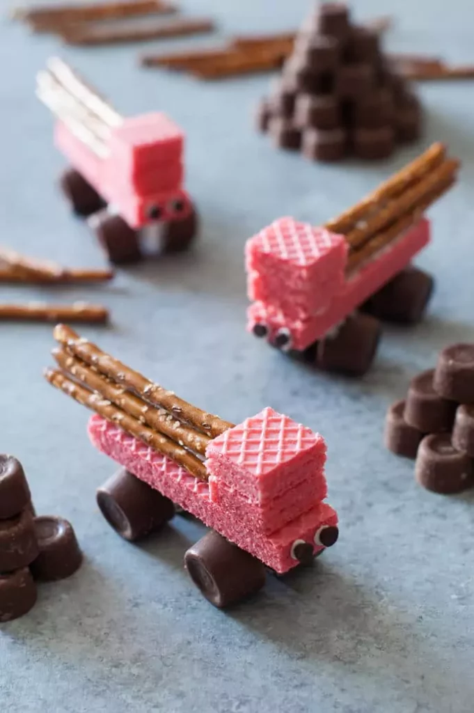 Creative edible crafts of trucks made from pink wafer cookies, pretzel sticks, and chocolate candies on a grey surface.
