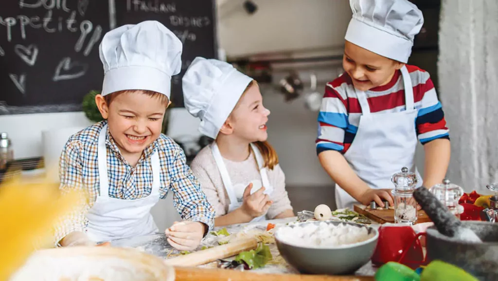 Three children wearing chef hats and aprons, engaging in a fun cooking activity surrounded by various ingredients, showcasing the joy of culinary learning and creativity. 