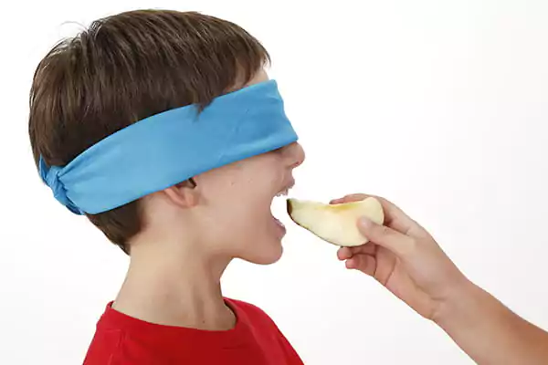 A kid with a blue blindfold is being handed an apple slice by another person.
