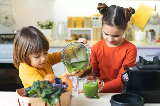 Two kids working together to make a green smoothie in a bright kitchen. One child pours the blended green mixture from a blender jug into a glass cup held by the other child. Fresh vegetables, including leafy greens and carrots, are visible in a basket on the counter. The setting is a well-lit, modern kitchen with various utensils.