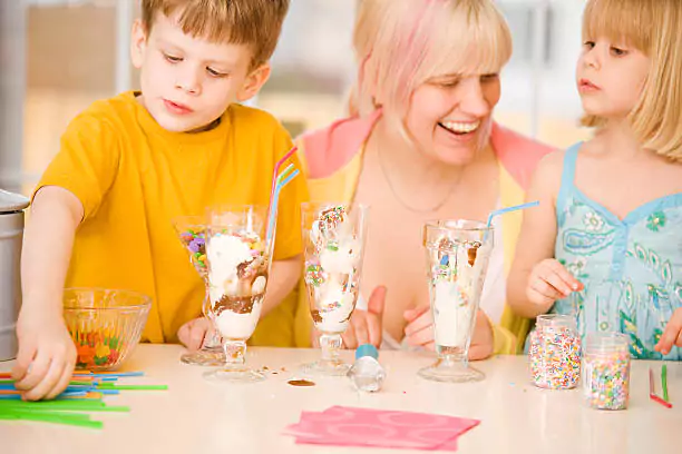 Children enjoying ice cream sundaes at a table with various toppings and utensils.
