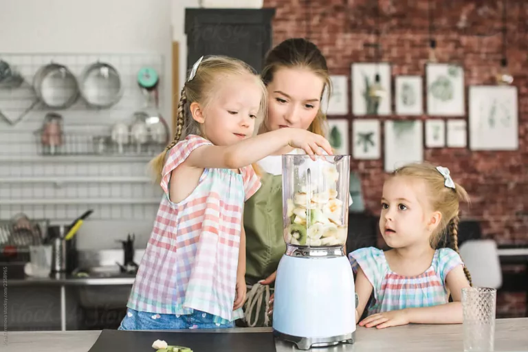 Three individuals with obscured faces are preparing a smoothie using a blender in a cozy, well-equipped kitchen. The scene captures a moment of collaboration in making a healthy snack, highlighting the homely atmosphere and teamwork.