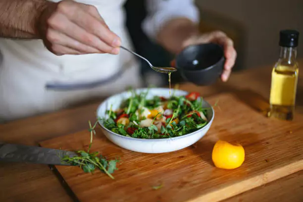A person drizzling dressing over a fresh salad, with ingredients and olive oil on a wooden table.
