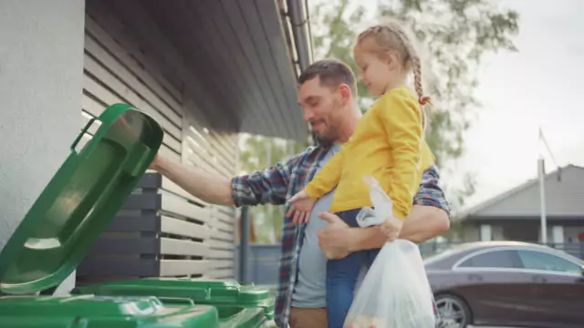 A person holding a child while disposing of trash into a green bin outside a building.
