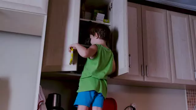 A child reaching into a kitchen cabinet while standing on a stool, depicting curiosity and exploration.
