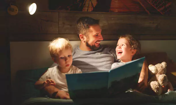 A person reading a book to two children, all seated on a couch under warm lighting, creating an intimate and cozy atmosphere.
