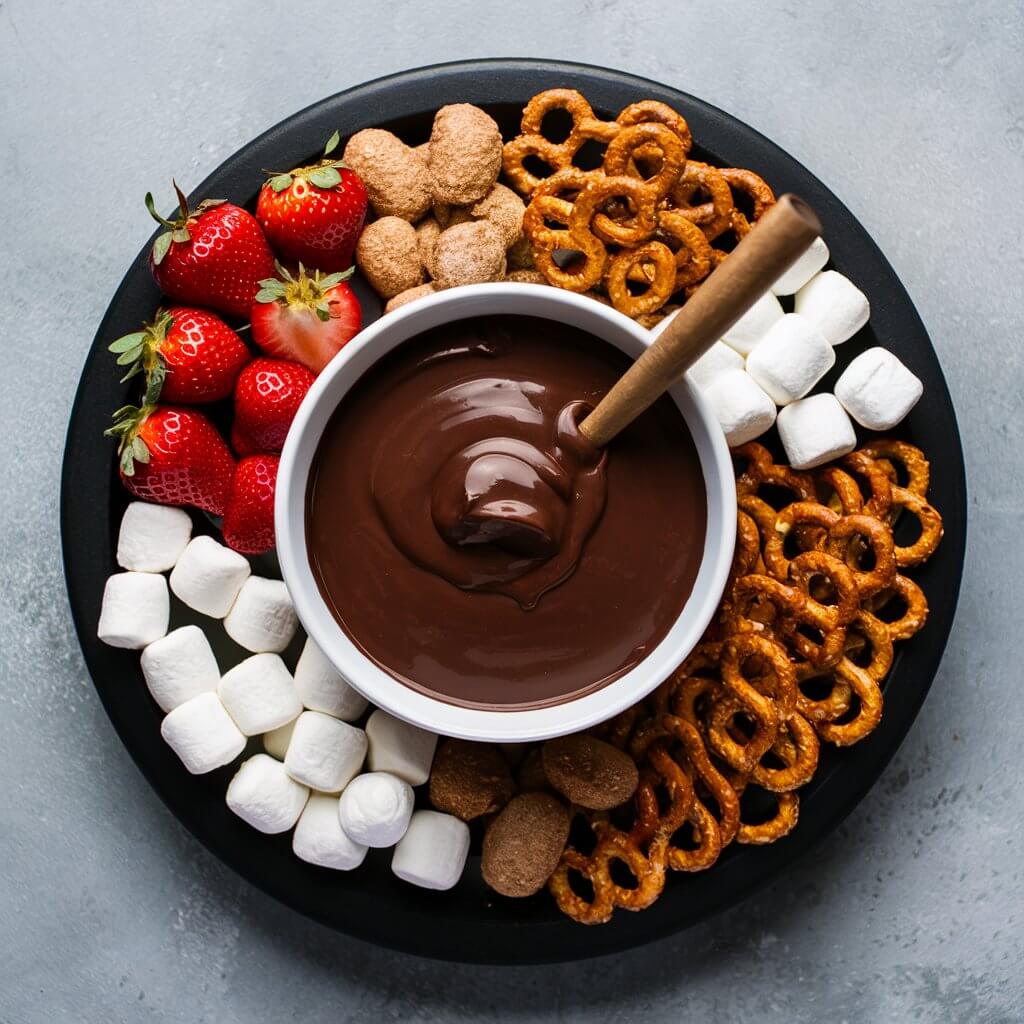 A platter with a chocolate fondue with an assortment of dippables such as strawberries, marshmallows, and pretzels arranged around it. The rich, chocolate creates a luxurious and engaging dessert experience.