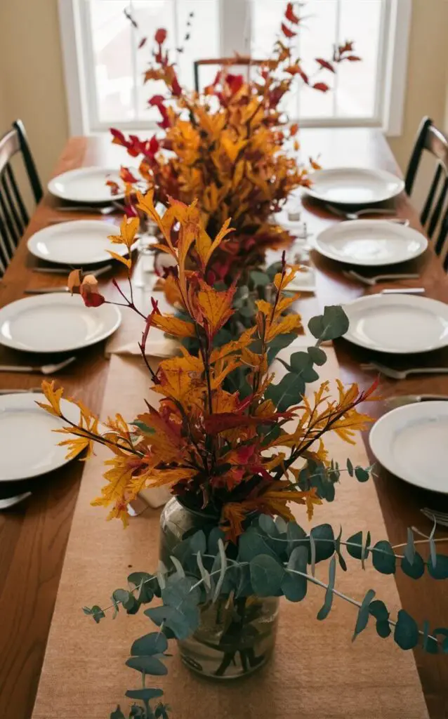 A dining table adorned with fall foliage as part of the decor. Branches with colorful leaves and eucalyptus are arranged in vases and laid directly on the table, bringing the beauty of the season indoors.