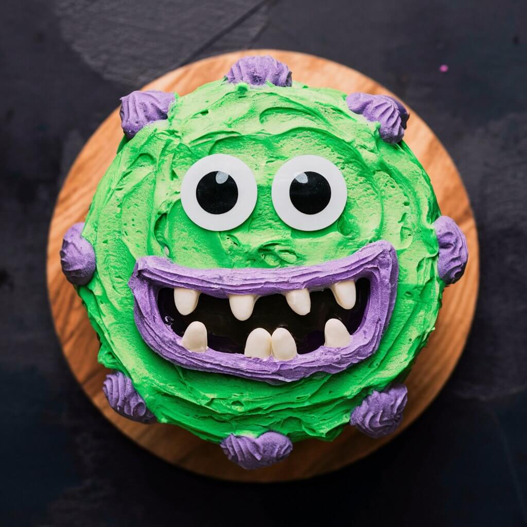 A brightly colored Halloween cake frosted in green and purple, adorned with edible googly eyes and candy teeth. The playful monster design includes various textures and shapes for added fun.