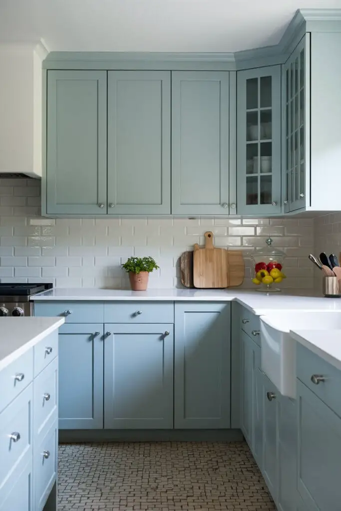 The photograph showcases a fresh and inviting kitchen characterized by a soothing powder blue and white color palette. The powder blue cabinets dominate the lower half of the kitchen, their soft hue providing a calm and cheerful atmosphere. These cabinets have sleek, modern lines, with minimalist silver handles that add a touch of sophistication without overwhelming the color scheme.

Above the cabinets, the countertops are pristine white, their smooth surface reflecting the ample natural light flooding into the space. The countertops are clutter-free, save for a few carefully chosen decorative items: a small potted plant in a ceramic pot, a wooden cutting board, and a glass jar filled with colorful fresh fruits.

The backsplash is composed of glossy white subway tiles, arranged in a classic horizontal pattern that adds texture and a bit of retro charm to the kitchen. The grout lines are thin and barely noticeable, ensuring the clean, seamless look of the backsplash. Above the backsplash, open shelves in the same powder blue color as the cabinets hold neatly organized white dishware and glassware, contributing to the airy, uncluttered feel.

The flooring is a light-colored wood, its natural grain providing a warm contrast to the cooler tones of blue and white. The kitchen is illuminated by a combination of natural light streaming through a large window above the sink and modern pendant lights hanging from the ceiling. These lights have simple, white shades that blend seamlessly with the overall color scheme.

In the center of the kitchen, a powder blue island with a white countertop offers additional workspace and storage. Around the island, a couple of white bar stools with powder blue cushions invite you to sit and enjoy a meal or a casual chat.

Overall, the kitchen feels bright, open, and welcoming, with its harmonious blend of powder blue and white creating a serene yet stylish environment.