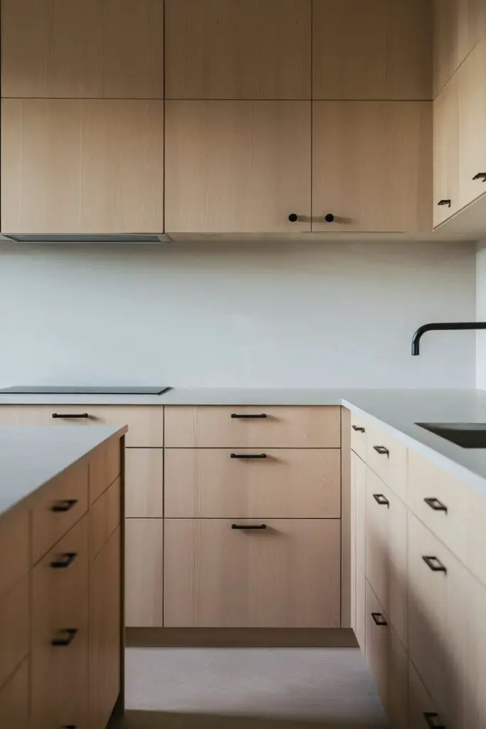 a close-up shot of of a Japandi kitchen, the design focuses on a harmonious blend of Japanese minimalism and Scandinavian simplicity. The kitchen space is characterized by its serene and uncluttered atmosphere.

The cabinets and drawers feature simple and sleek hardware, emphasizing minimalist handles and knobs. These fixtures are either in matte black or brushed nickel, contributing to the clean aesthetic of the kitchen. The matte black finish provides a striking contrast against the light-colored cabinetry, while the brushed nickel option adds a subtle sheen that complements the overall muted tones.

The cabinetry itself is likely made of light-colored wood or laminate, showcasing a smooth and seamless surface without elaborate detailing. The countertops appear to be crafted from a durable material like quartz or light-colored stone, adding a touch of elegance to the understated design.

The kitchen layout is functional and efficient, with ample storage concealed within the cabinets. Open shelving might be integrated sparingly to display a few carefully selected items, maintaining the minimalist theme.

Natural light filters into the space, enhancing the warmth of the wooden elements and creating a welcoming ambiance. The flooring is likely to be hardwood or a light-toned tile, further contributing to the airy and spacious feel of the room.

Overall, the Japandi kitchen exudes a sense of tranquility and balance, combining the timeless appeal of Japanese and Scandinavian design principles into a modern and inviting culinary space.