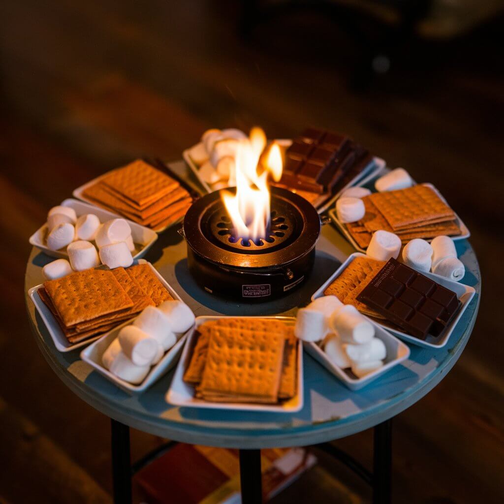 A cozy, inviting scene of a s'mores bar setup on a small wooden table. The table is adorned with neatly arranged graham crackers, assorted chocolate bars, and fluffy marshmallows. In the center, a small tabletop burner with a flickering flame provides the perfect space for guests to toast their marshmallows. The warm glow from the flame creates a nostalgic, campfire-like ambiance, inviting friends to gather around and create their own delicious s'mores masterpieces.