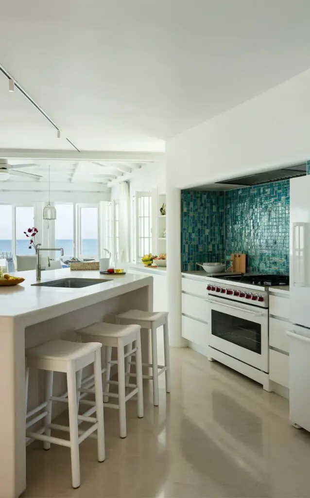 A pristine, modern coastal kitchen with sleek white appliances and minimalist design. The space features a large island with white stools, a stainless steel sink, and a white oven and refrigerator. The backsplash is a stunning mosaic of blue and green tiles, evoking the feeling of an ocean breeze. The room is filled with natural light from large windows, and there's a beautiful view of the sea in the distance.