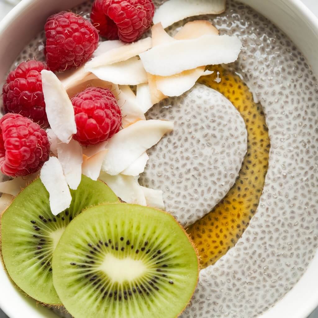 A photo of a bowl of chia seed porridge. The porridge has a speckled, pudding-like texture due to the tiny black chia seeds. The bowl is topped with coconut flakes, fresh raspberries, and a drizzle of honey. The glossy chia seeds and the bright green sliced kiwi add a tropical, refreshing look to the dish.