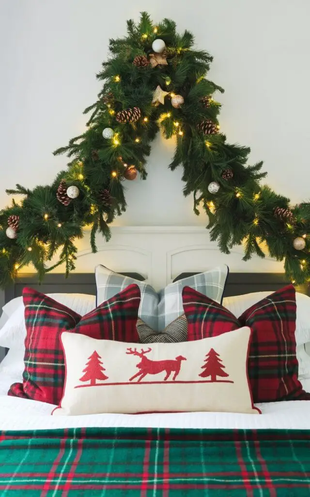 A cozy Christmas bedroom with a lush green garland draped over the headboard. The garland is adorned with small ornaments, pinecones, and twinkling lights, creating a festive focal point. The bed features a plaid blanket and holiday-themed pillows, adding to the seasonal charm.