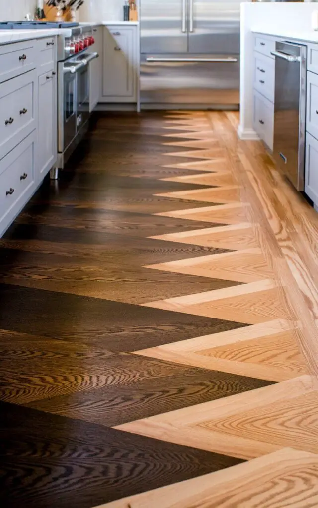 A photo of a kitchen with a dynamic floor made of hardwood planks. The floor has a gradient from dark espresso to light oak, with a smooth transition between the two colors. This creates a striking visual effect, adding depth and dimension to the room. The light gray cabinets and stainless steel appliances complement the bold floor, making it the focal point of the kitchen's design.