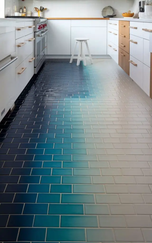 A photo of a kitchen with a mesmerizing gradient floor. The floor tiles transition from dark navy blue at the back of the kitchen to soft aqua at the front. The gradient effect creates a flowing, dynamic look, mimicking the movement of water. The kitchen has white cabinets and light wood accents. There is a small white stool near the floor. The overall ambiance of the kitchen is fresh and modern, with a hint of coastal inspiration.