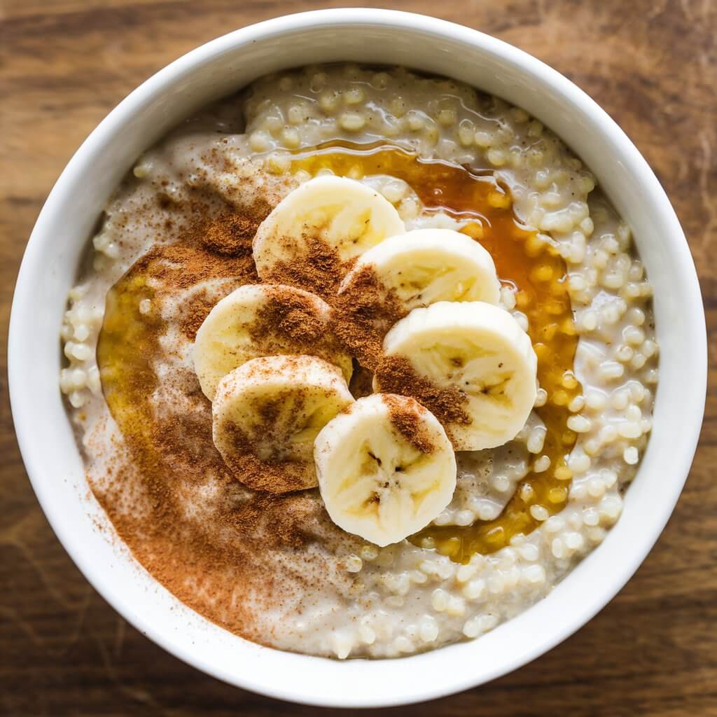 A photo of a bowl of quinoa porridge topped with sliced bananas, a sprinkle of cinnamon, and a drizzle of maple syrup. The quinoa grains are cooked to a soft, fluffy consistency, and the bright yellow bananas and rich golden syrup add a touch of sweetness to the hearty quinoa base. The bowl is placed on a wooden surface.