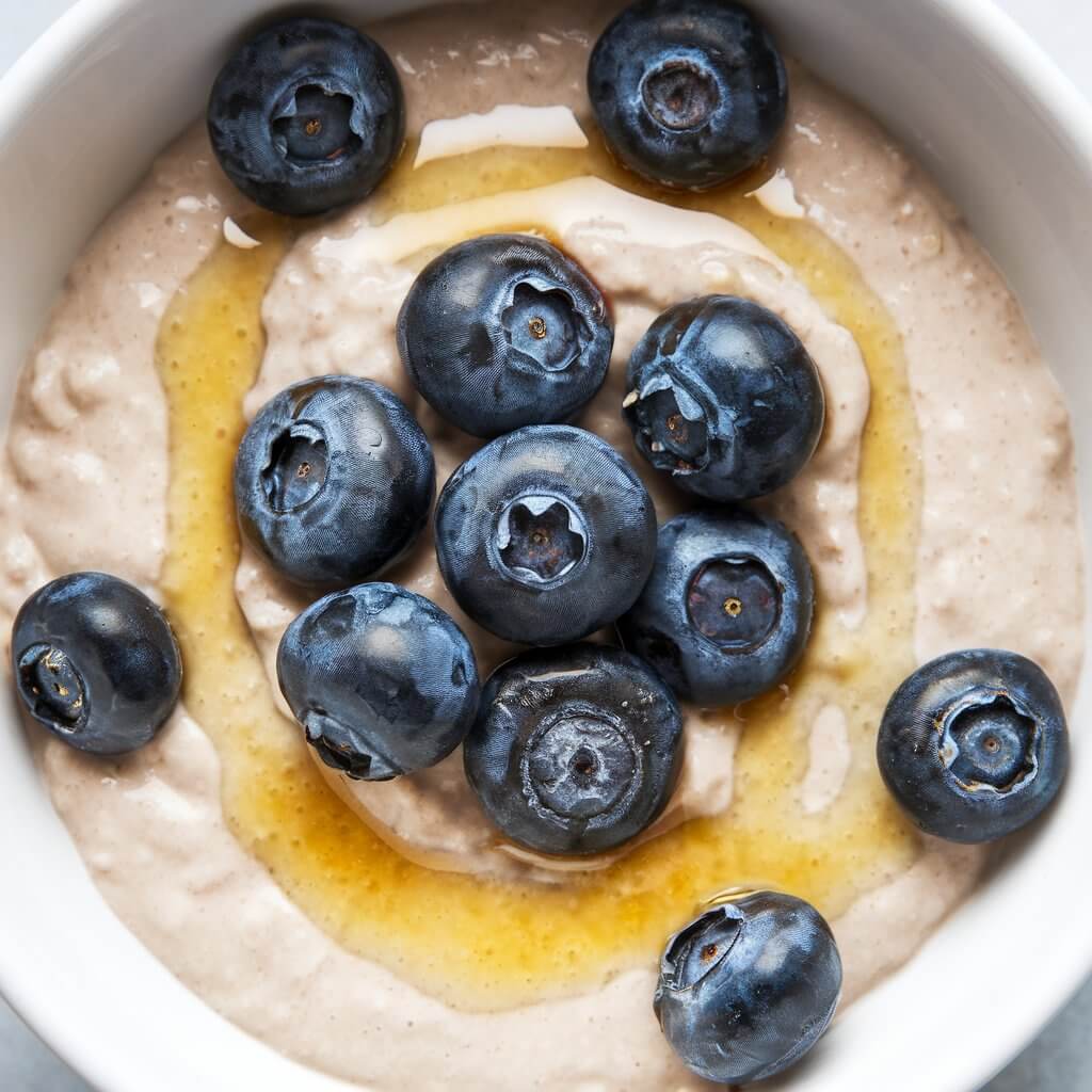 A photo of a bowl of vanilla and honey porridge. The porridge has a creamy, smooth texture and is infused with vanilla extract. It is topped with a drizzle of golden honey and fresh blueberries. The pale, creamy base of the porridge is beautifully complemented by the bright honey and berries. The background is simple and clean, allowing the porridge to be the main focus of the image.