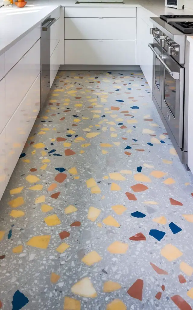 A photo of a kitchen with a vibrant terrazzo floor. The floor contains a mix of colorful marble chips in shades of yellow, blue, and coral, set against a light gray background. The terrazzo adds a playful pop of color to the minimalist kitchen, which features white countertops and stainless steel appliances. Natural light is bouncing off the terrazzo surface, illuminating the space.