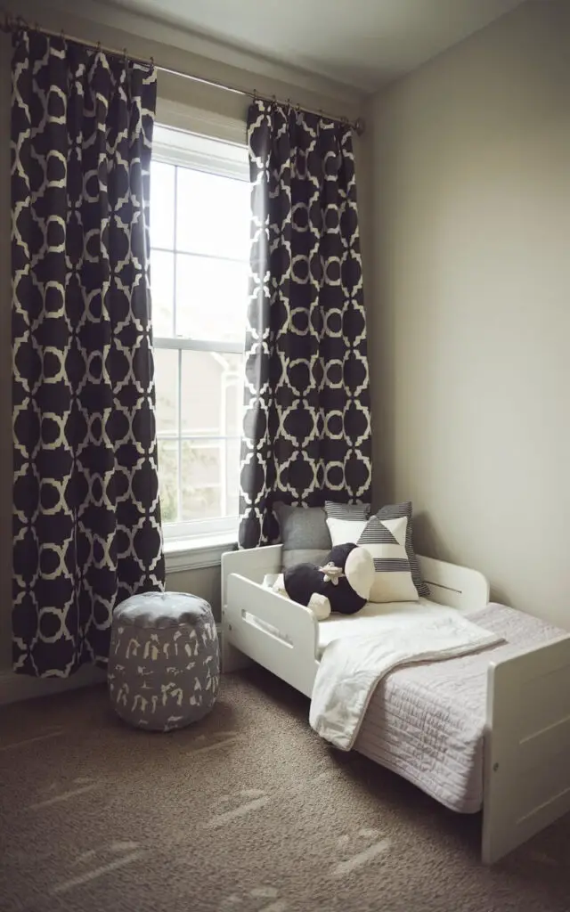 A small toddler bedroom with a dark, patterned blackout curtain. The cozy toddler bed is positioned next to the window. The curtain is drawn to block light for daytime naps or early bedtime routines, providing both style and functionality.