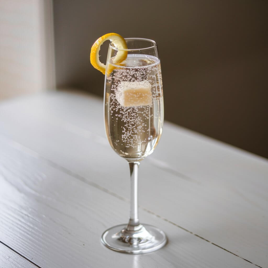 A photo of a classic champagne cocktail in a tall flute. The flute is filled with sparkling bubbles and has a sugar cube at the bottom and a twist of lemon on top. The glass is set against a bright wooden white table.