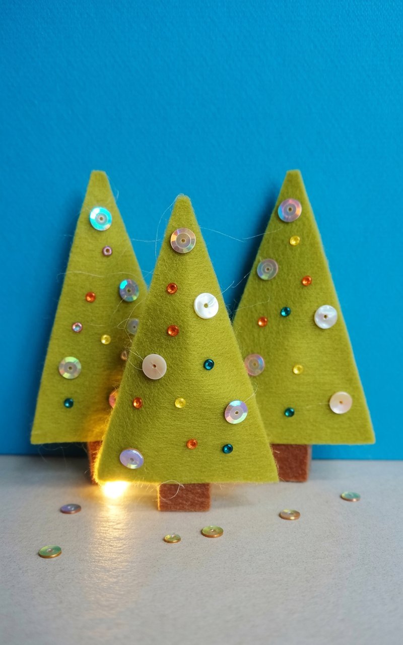 Several miniature felt Christmas trees stand on a smooth white surface, each approximately six inches tall. The trees are decorated with iridescent sequins, small glass beads in various colors, and tiny, round buttons made of mother-of-pearl. Scattered around the base of the trees are loose sequins and beads, adding a sense of whimsical detail. A soft glow illuminates the scene, creating a warm ambiance and subtle shadows beneath the trees.